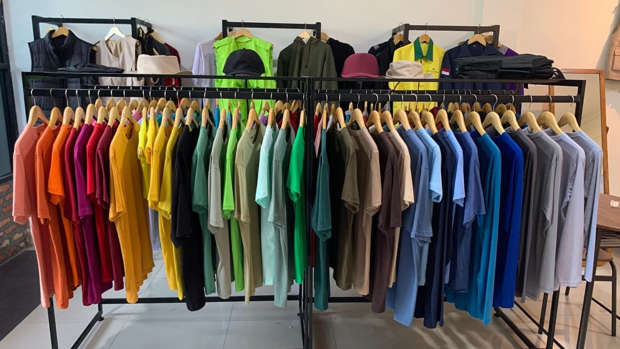 Rak kaos polos warna-warni beserta contoh topi, rompi, dan seragam di showroom